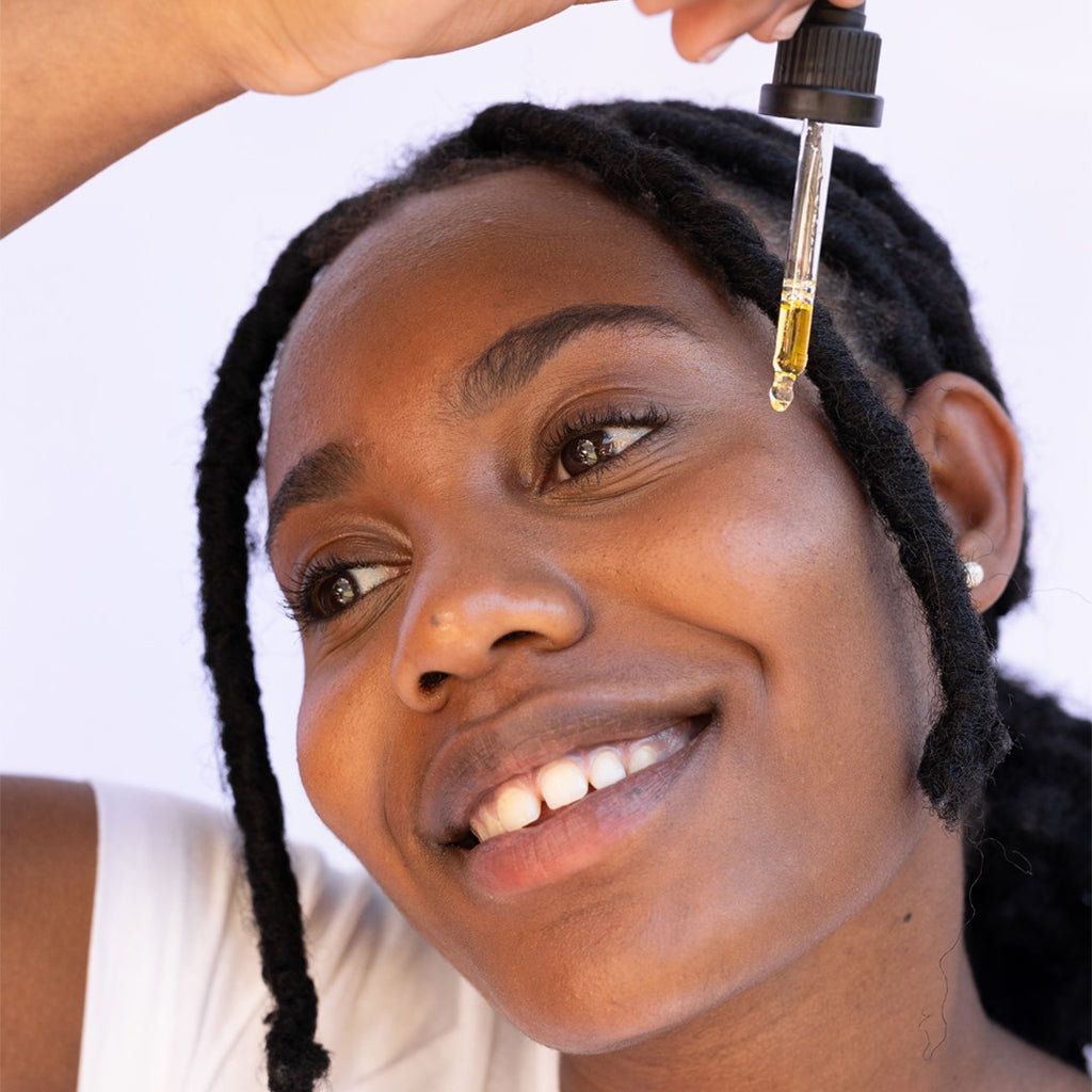 Femme noir souriante qui tient une pipette avec une goutte de sérum soin visage prête à tomber 