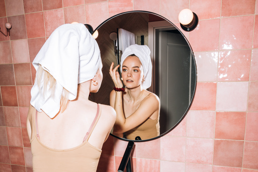 femme portant un débardeur de couleur clair et avec une serviette enroulée sur sa tête se regarde dans un miroir