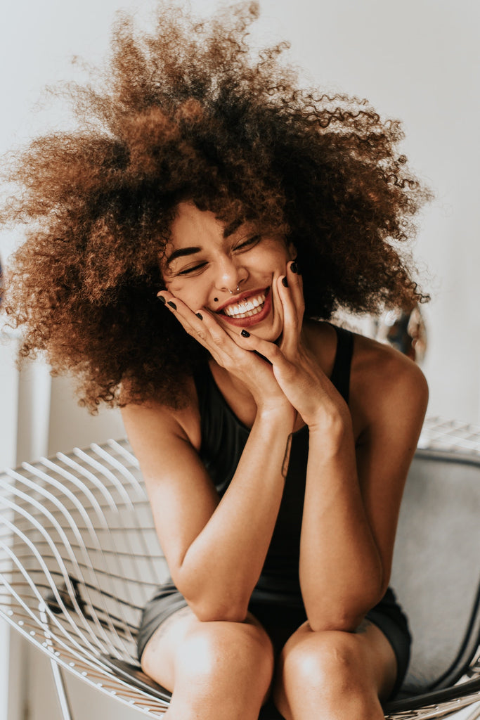 femme est assise sur un fauteuil blanc, elle se tient le visage avec ses deux mains. Elle a un grand sourire et des cheveux texturés