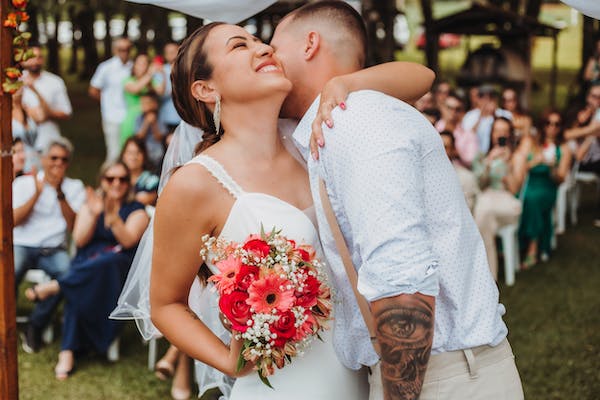 Deux jeunes mariés se prennent dans les bras, la mariée a un très beau bouquet de fleurs rouges et roses.